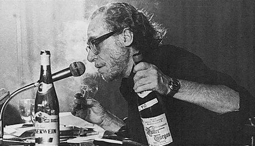 Photo of Charles Bukowski sitting at a desk covered in papers, speaking into a microphone, holding a cigarette in one hand and a bottle of alcohol in the other. 